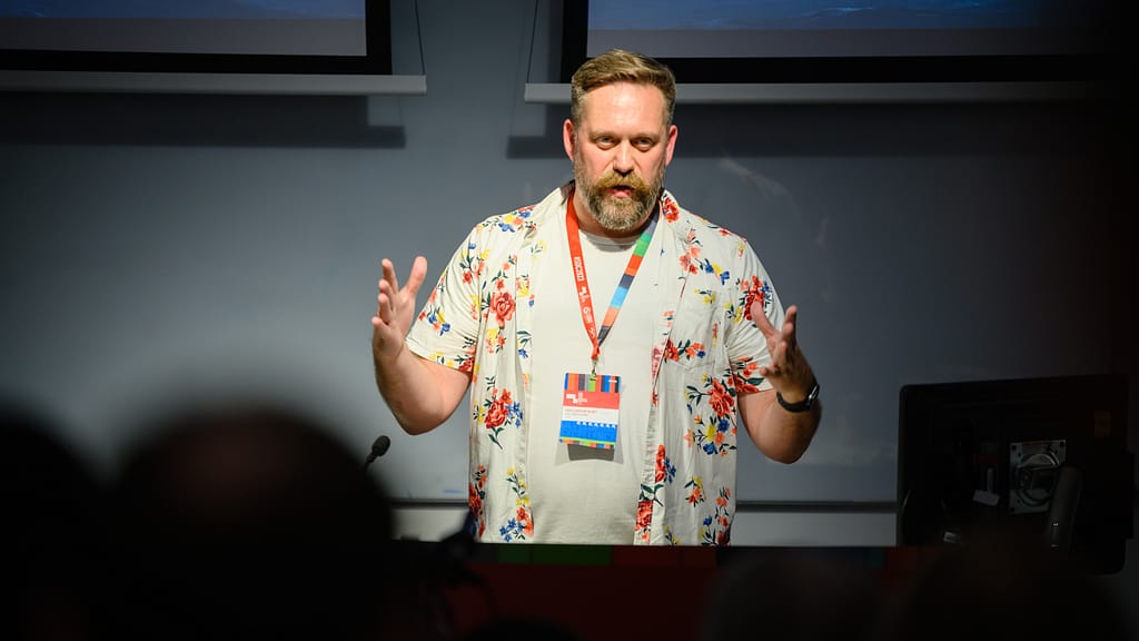 Louis Larsson-De Wet speaking at a conference. The photo is taken from the crowd and he is lit by a spotlight.